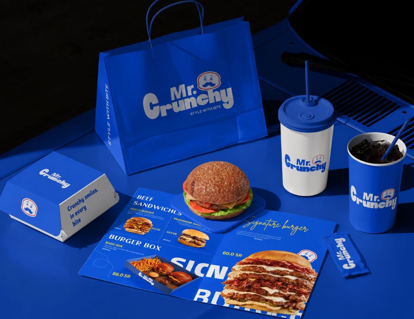 Mr. Crunchy branded packaging with blue takeaway bag, burger box, fries, drink, and sauces.